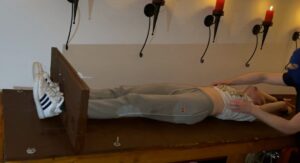 Woman restrained on a table with her arms extended, wearing casual attire and sneakers, in a room wi.