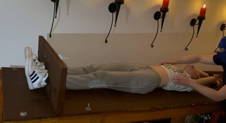 Woman restrained on a table with her arms extended, wearing casual attire and sneakers, in a room wi.