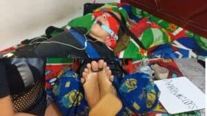 A woman is restrained on a bed, her feet held by someone else, wearing glasses and casual clothing.