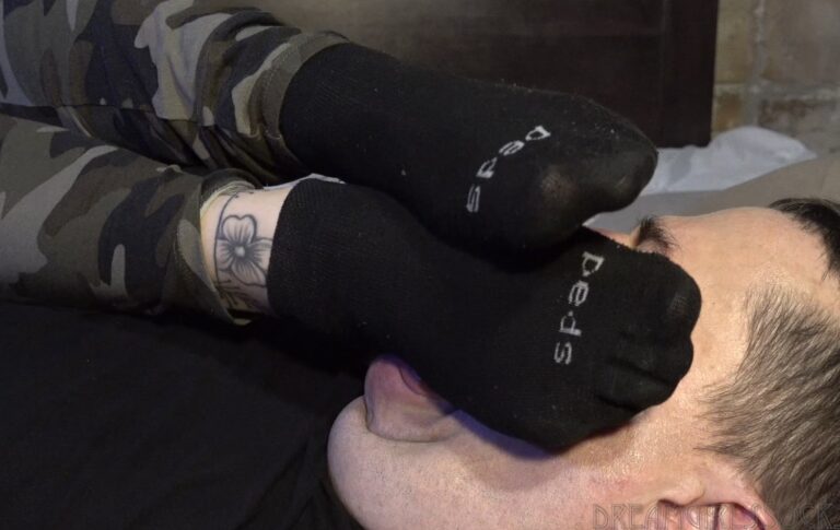 Woman in camouflage pants and black gloves tickling a man lying face down.