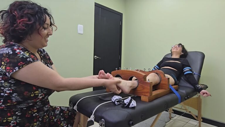 Woman in black outfit being tickled on her feet with her hands restrained.