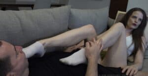 Woman restrained, being tickled on sofa, with long brown hair and casual outfit.