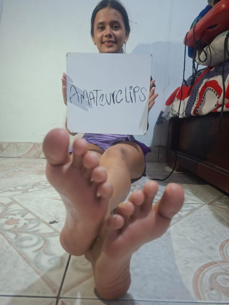 Girl with dark hair, sitting on the floor, holding a sign that says "Amateurclips," with her feet pr.