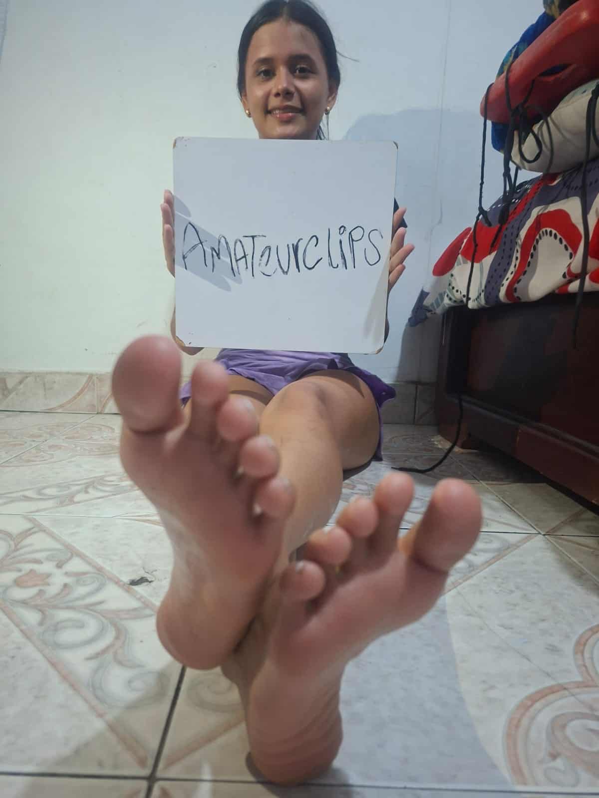 Girl with dark hair, sitting on the floor, holding a sign that says "Amateurclips," with her feet pr.